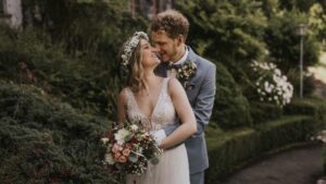 Ein Brautpaar lächelt sich an, umgeben von üppigem Grün und Blumen, während sie in eleganter Hochzeitskleidung posieren.