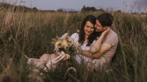 Ein Paar sitzt im hohen Gras, umarmt sich und lächelt, während es einen Blumenstrauss in der Hand hält.