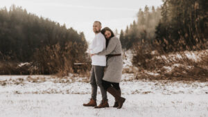 Paar umarmt sich in einer verschneiten Landschaft, umgeben von Bäumen und trockenem Gras im sanften Winterlicht.