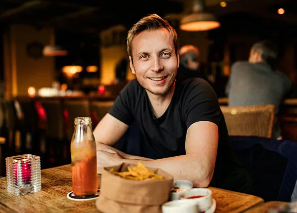 STARTSEITE 1 Mann sitzt entspannt an einem Tisch in einer Bar, geniesst ein Getränk und Snacks in gemütlicher Atmosphäre.