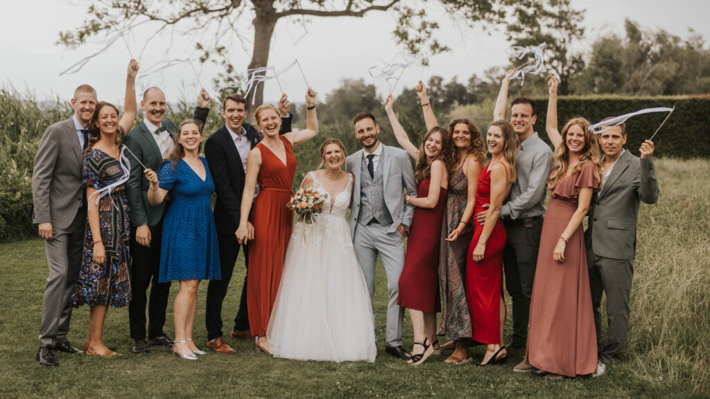 Hochzeitsgesellschaft feiert im Freien mit Luftschlangen, umgeben von grünen Wiesen und Bäumen. Braut strahlt im eleganten Kleid.