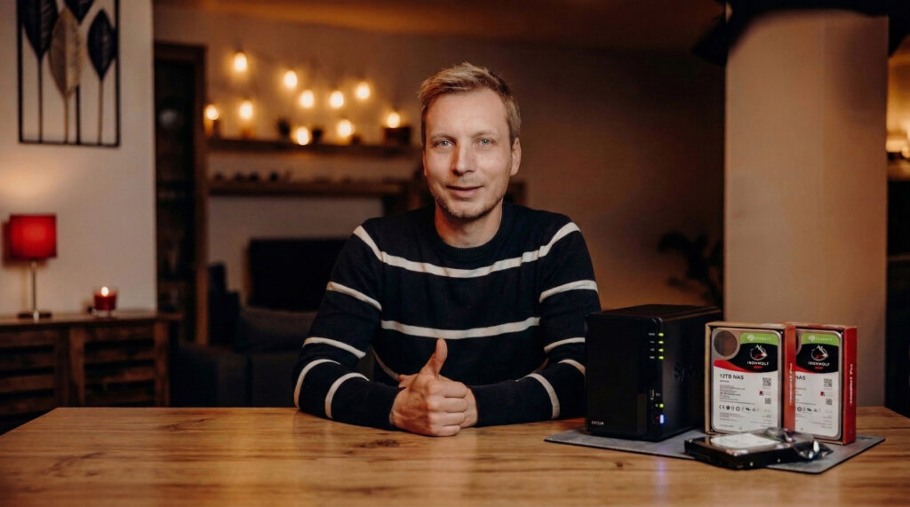 Mann gibt Daumen hoch und präsentiert NAS-Gerät sowie Festplatten auf einem Holztablett in gemütlicher, beleuchteter Umgebung.