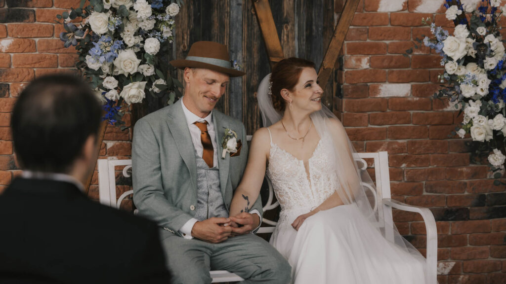 Brautpaar sitzt gemeinsam auf einer Bank, umgeben von Blumenarrangements, während sie sich an den Händen halten.