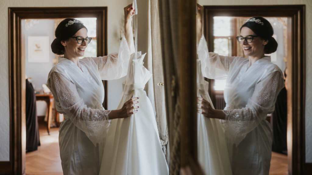 Die Braut hält ihr Hochzeitskleid in einem eleganten Raum und betrachtet sich lächelnd im Spiegel.