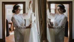 Die Braut hält ihr Hochzeitskleid in einem eleganten Raum und betrachtet sich lächelnd im Spiegel.