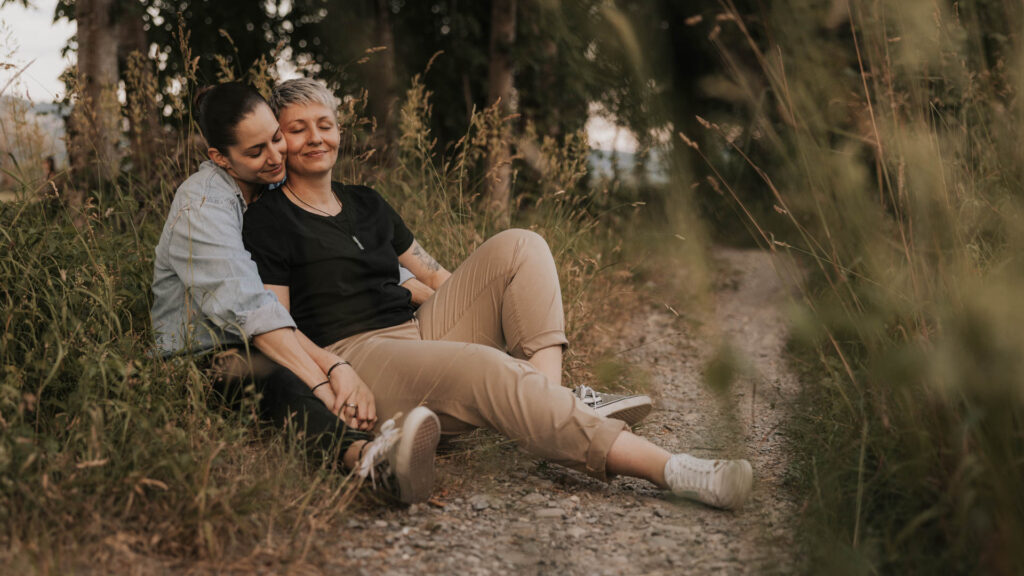 Zwei Frauen sitzen entspannt auf einem Kiesweg, umgeben von hohem Gras, und halten sich liebevoll an den Händen.