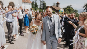 Braut und Bräutigam gehen lächelnd Hand in Hand durch eine jubelnde Gästeschar in malerischer Umgebung.